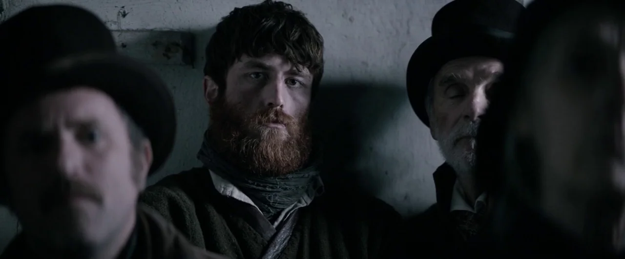 Black ’47 (2018) movie still: medium — Three men sit shoulder-to-shoulder against a grey, textured stone wall. They wear 19th-century attire…