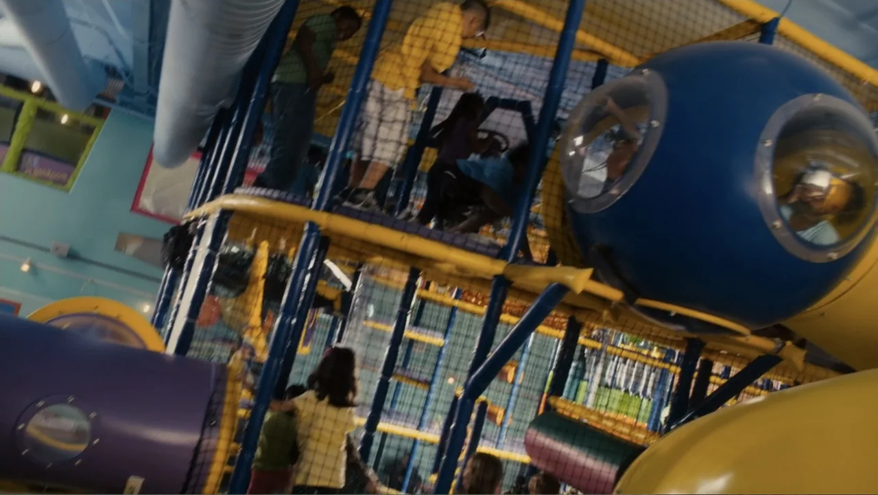 Just Go with It (2011) movie still: medium — A multi-level indoor playground constructed with blue metal poles and yellow plastic tunnels. Multiple…