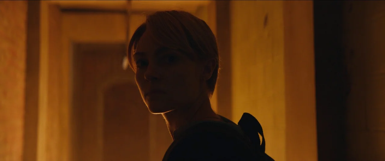 Rebel Ridge (2024) movie still: closeup — A dimly lit narrow corridor with orange light spilling from one direction. A young woman with short blonde…