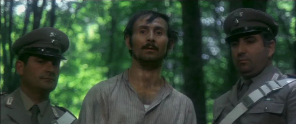 Don’t Torture A Duckling (1972) movie still: medium — Two uniformed soldiers stand flanking a man in a light striped shirt. The group is positioned…