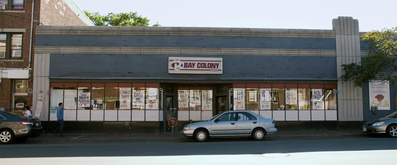 Ted 2 (2015) movie still: wide — A single-story commercial building with a grey brick facade and large display windows. A man walks on the sidewalk…