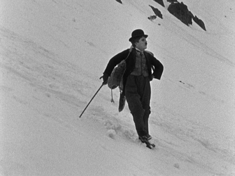 The Gold Rush (1925) movie still: fullbody — A snow-covered mountainside slopes downwards with dark rocky outcroppings visible in the upper right. A…
