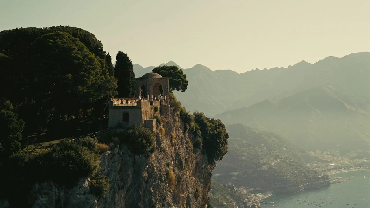 Tenet (2020) movie still: establishing — A stone gazebo sits atop a rugged cliff overlooking a mountain range and sea. Dense green foliage surrounds…