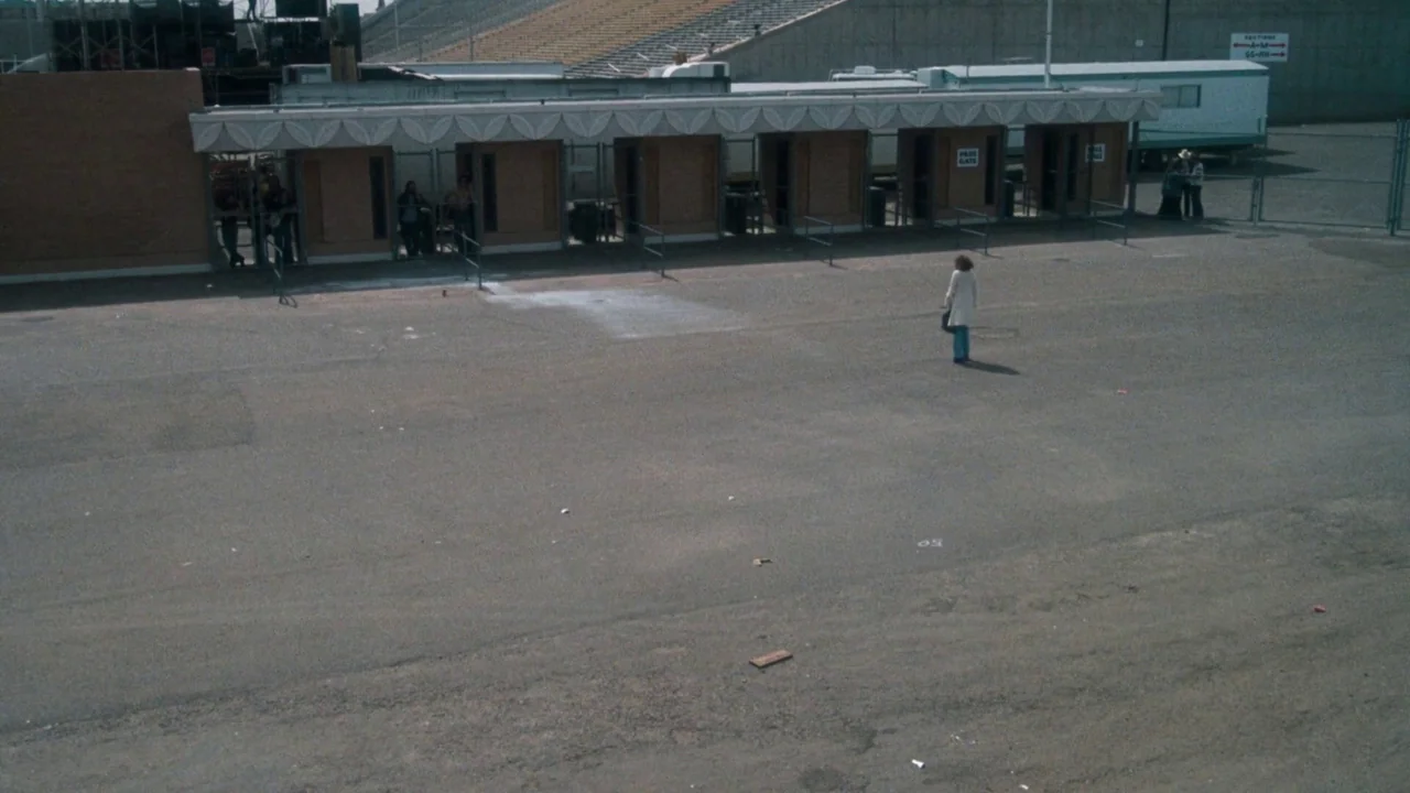 A Star Is Born (1976) movie still: wide — A vast, desolate asphalt lot sits before a row of stadium ticket booths. A woman in a light coat stands…