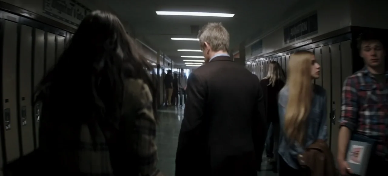 I Still See You (2018) movie still: medium — A school hallway is lined with grey metal lockers on both sides. A man in a dark suit walks forward…