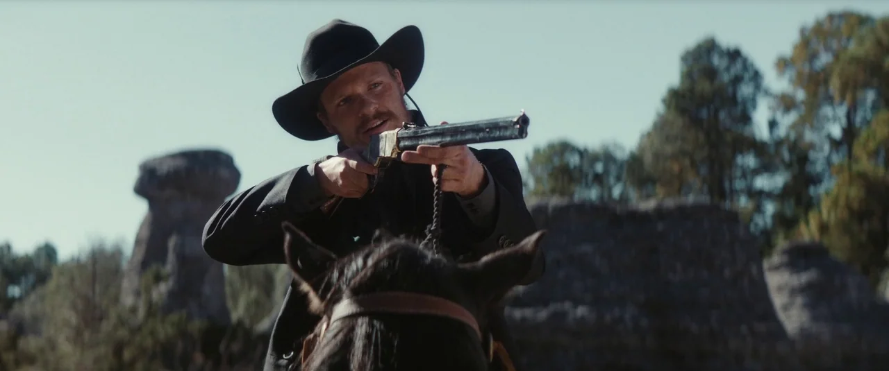 The Dead Don’t Hurt (2023) movie still: medium — A man rides a horse through a barren landscape featuring tall, eroded rock formations. The man…