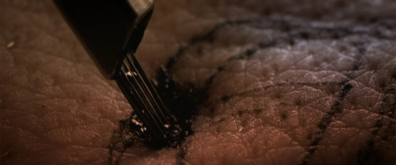 Final Destination Bloodlines (2025) movie still: closeup — A close-up view reveals human skin being marked by a metal tattoo needle. Thick black ink…