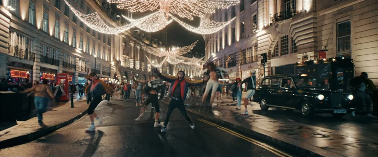 Better Man (2024) movie still: wide — A bustling city street at night is decorated with overhead holiday lights and traffic. Several individuals are…