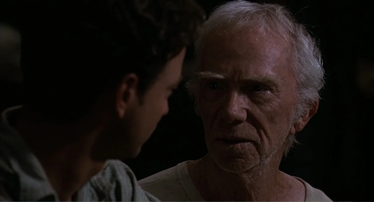 Of Mice and Men (1992) movie still: medium — A dark indoor space with minimal ambient light and deep shadows. An older man with grey hair engages in…
