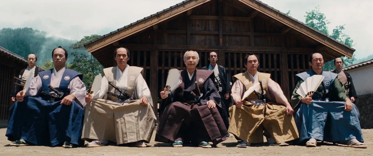 Silence (2016) movie still: fullbody — Several men in traditional Japanese clothing sit in a line before a wooden building. They are positioned…