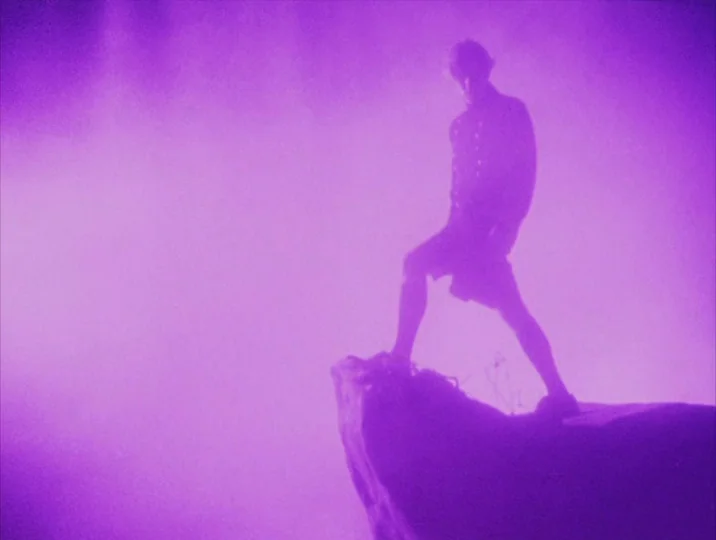 Careful (1992) movie still: fullbody — A figure stands on a rocky outcrop surrounded by thick, vibrant violet mist. The subject is backlit and…