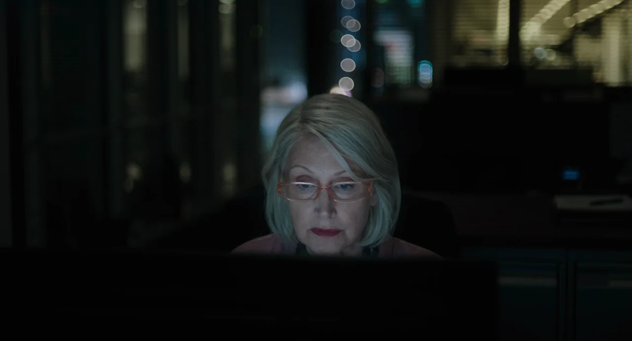 She Said (2022) movie still: closeup — A dark office environment at night features rows of desks and glass partitions. An older woman with silver…
