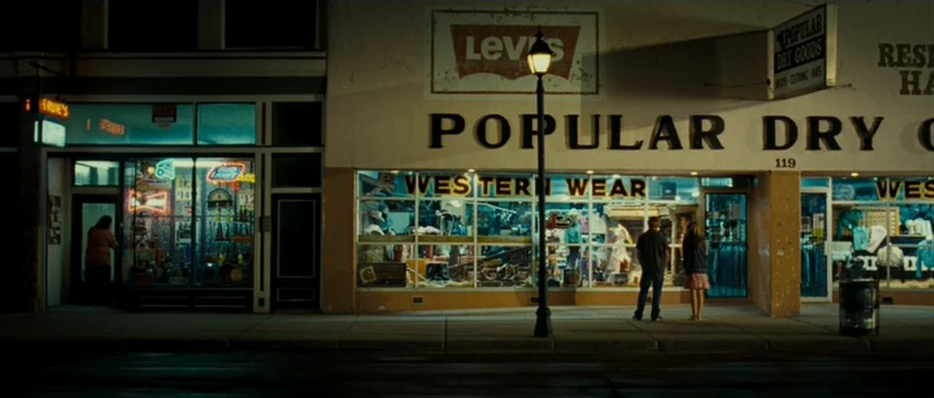 Paul (2011) movie still: wide — A quiet sidewalk at night featuring storefronts with glowing windows and signage. A man and a woman stand…