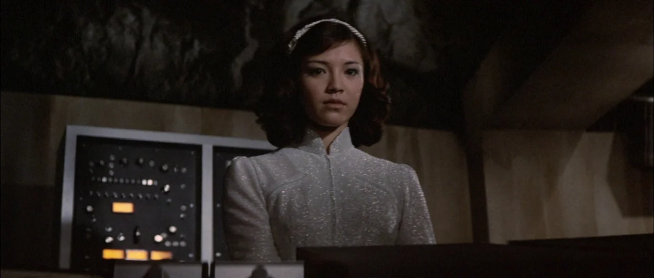 Terror of Mechagodzilla (1975) movie still: medium — A dimly lit room features a large technical console with illuminated control panels. A young…
