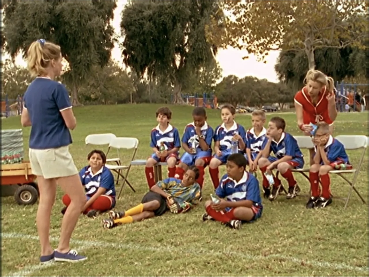 Just 4 Kicks (2003) movie still: fullbody — A sunny public park area with trees and a mowed grass field. A woman in a blue polo stands near a small…
