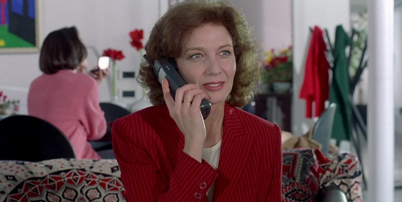 The Flower of My Secret (1995) movie still: medium — A modern living room with a patterned sofa and abstract artwork on the wall. A woman talks on a…