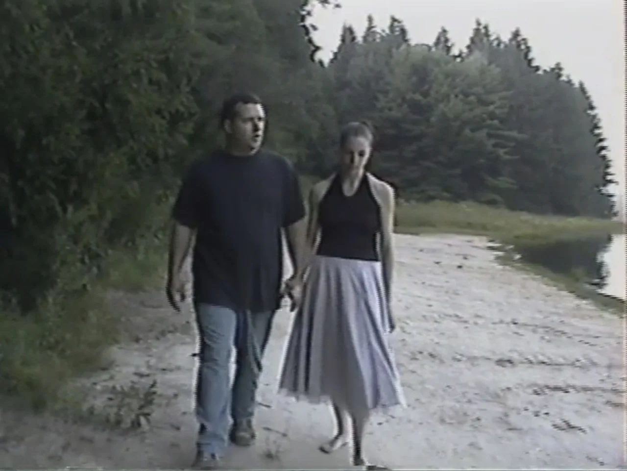 Dracula, Prisoner of Frankenstein (1972) movie still: fullbody — A man and a woman walk along a sandy lakeshore lined with pine trees. They traverse…