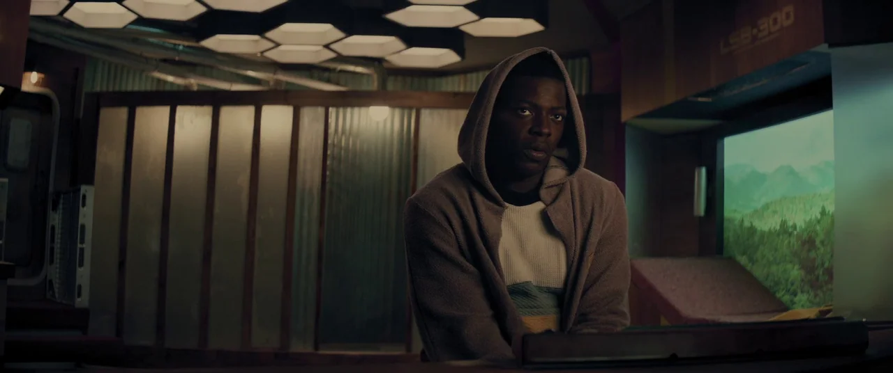 Domain (2017) movie still: medium — A dimly lit industrial room features hexagonal light panels mounted on the ceiling above. A man in a hooded…