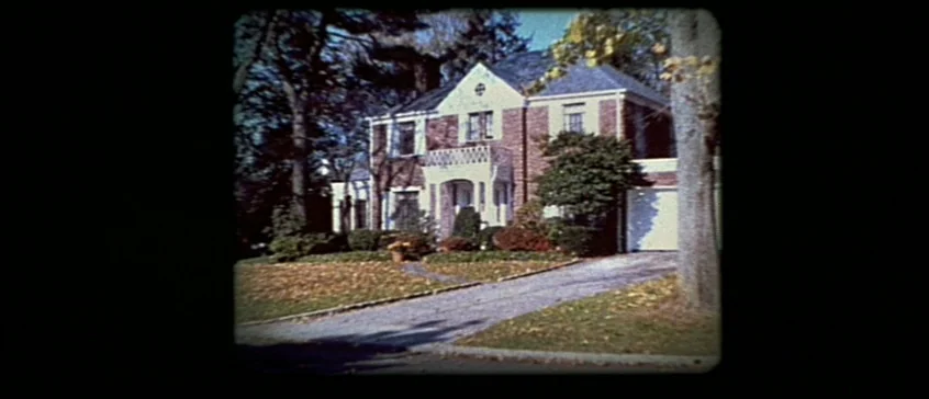 Man On The Moon (1999) movie still: establishing — A two-story suburban brick house with a white garage sits at the end of a driveway. Mature trees…