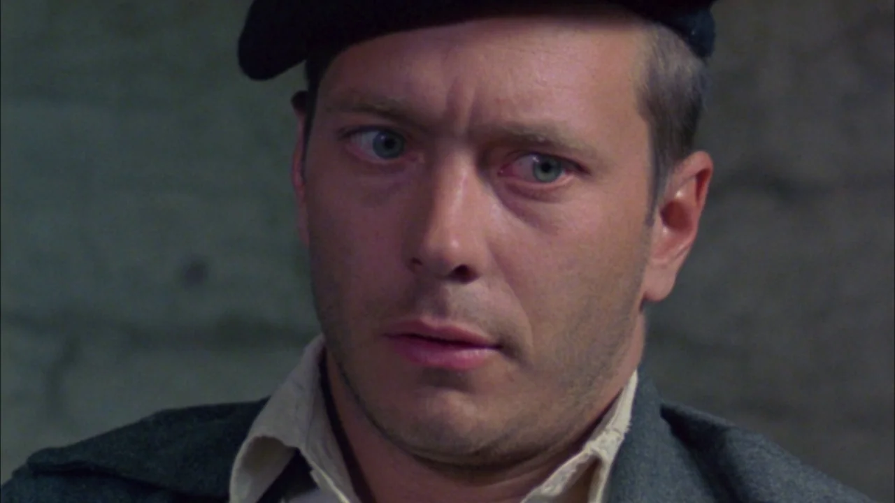 Poison movie still: closeup — A dark, dimly lit bunker room with rough, gray stone walls in the background. A man wears a dark beret and military…