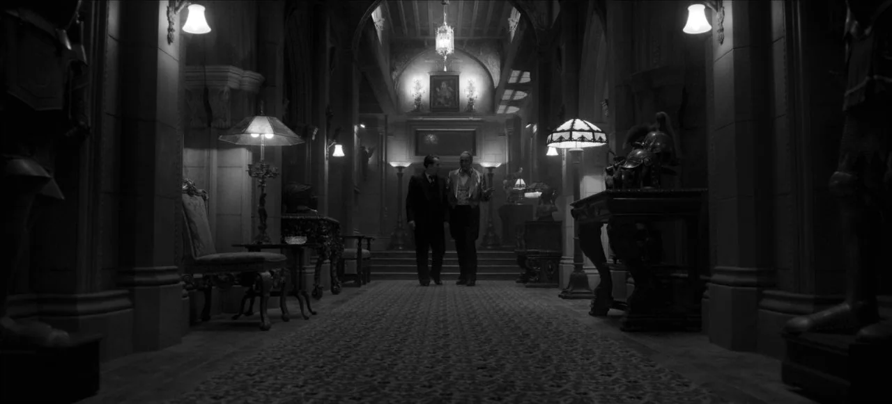 Mank (2020) movie still: wide — A dark, expansive mansion foyer with arched doorways and multiple glowing table lamps. Two formally dressed men walk…