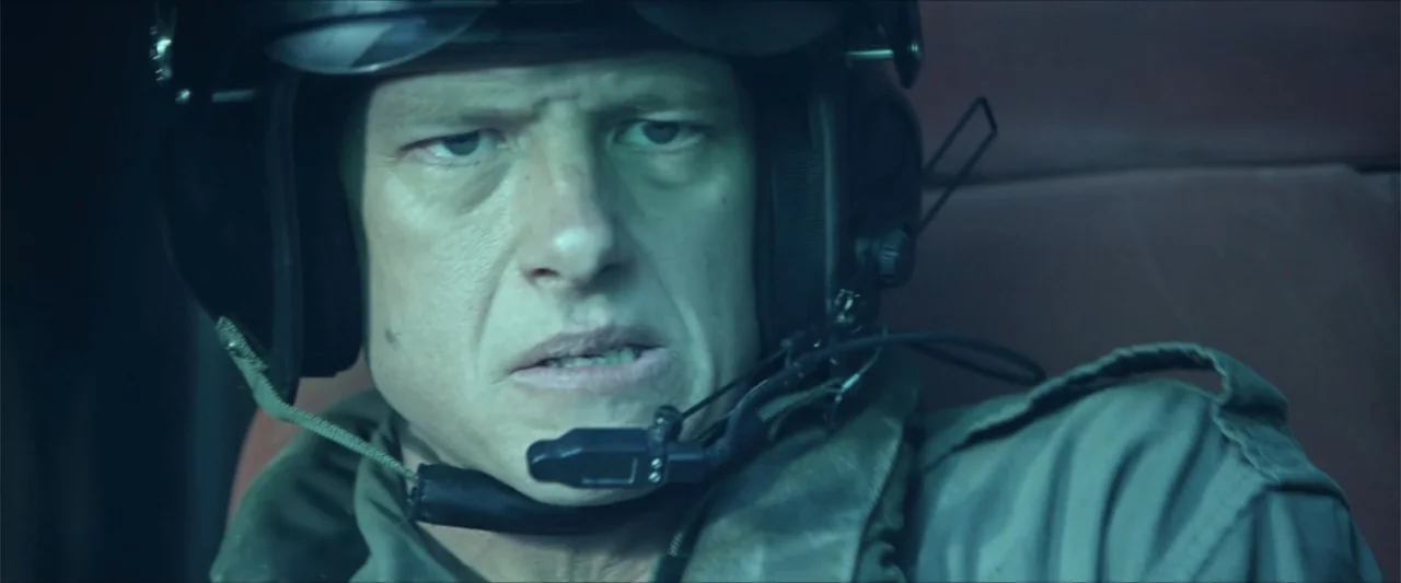Monster Hunter (2020) movie still: closeup — A close view inside a dimly lit helicopter cockpit with metallic textures. A man in a flight helmet…