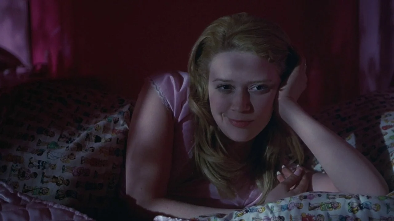 But I’m A Cheerleader (1999) movie still: medium — A dimly lit room features a bed covered in patterned cartoon-print linens. A woman with blonde…