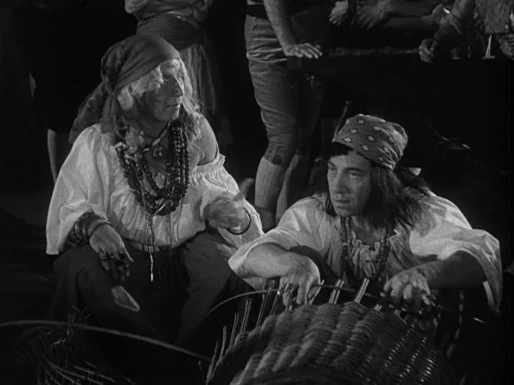 A Night at the Opera (1935) movie still: medium — Two individuals sit on the ground in a dimly lit, cluttered workspace. They are engaged in weaving…
