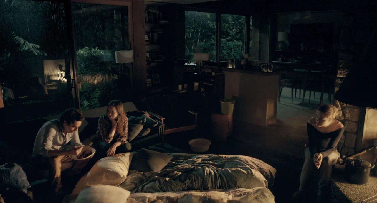 En lo profundo del bosque movie still: wide — A dimly lit room with a fireplace and large windows showing a rainy night. Two people sit on a couch…