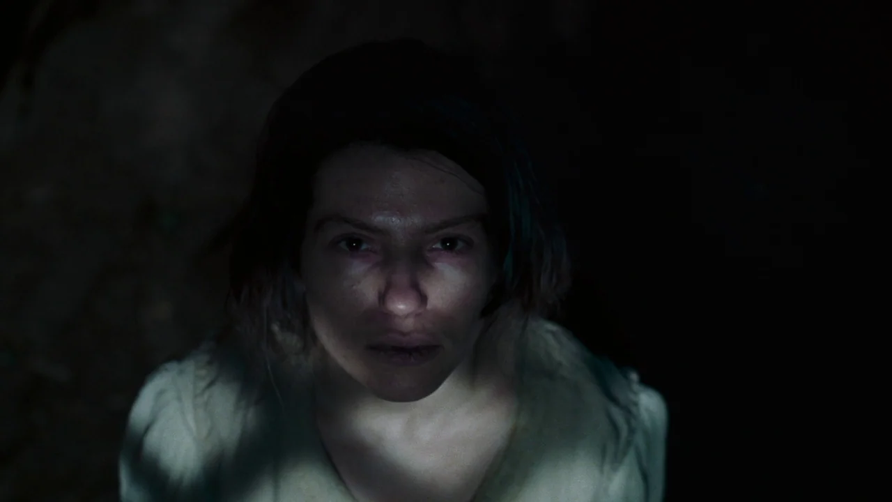 The Devil’s Bath (2024) movie still: closeup — A dark, underexposed interior space surrounds the subject. A woman faces the camera while lit from…