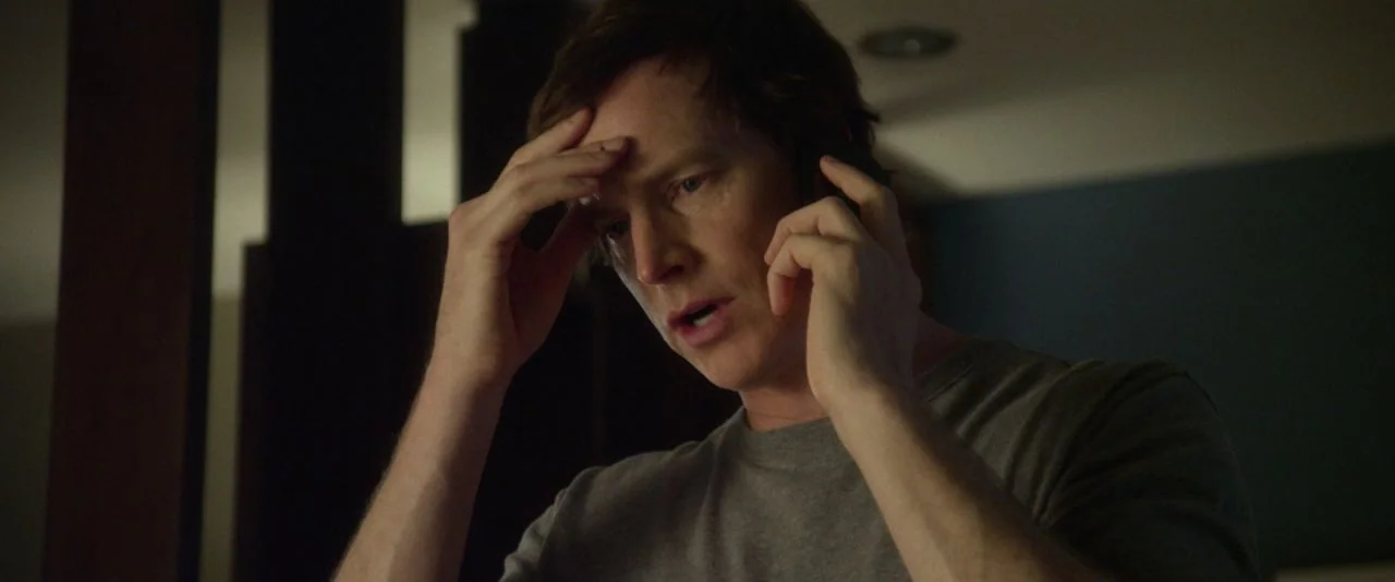 Night Owls (2015) movie still: medium — A dimly lit residential interior features a person in a plain gray t-shirt. The man holds his hand to his…
