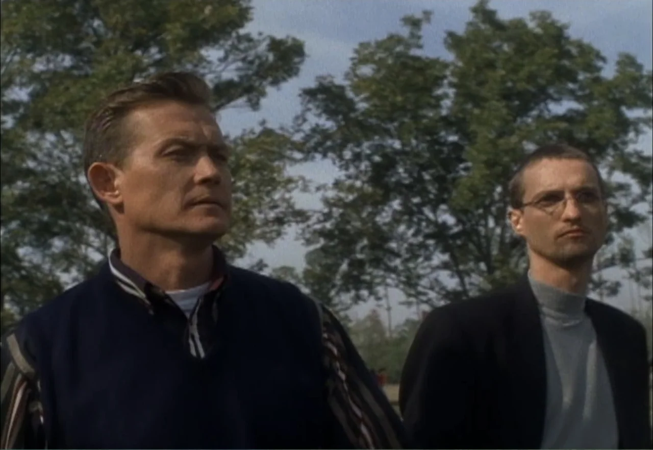 Ambushed (1998) movie still: medium — A green park with tall deciduous trees stands behind two men outdoors. The two men stand near each other…