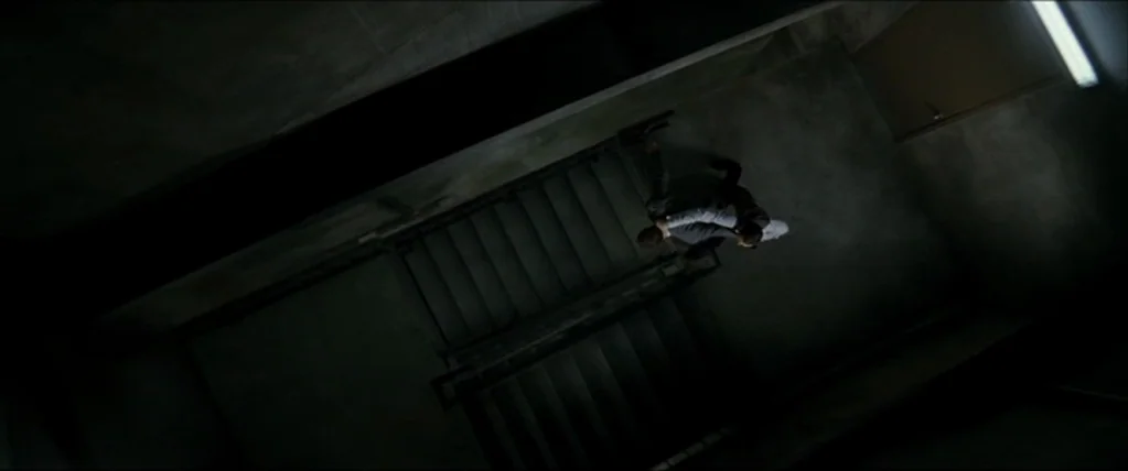 Inception (2010) movie still: wide — A concrete stairwell features stark walls and a metal railing under dim fluorescent light. Two men struggle…