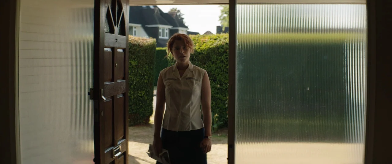 Beast (2017) movie still: medium — A woman stands in the open doorway of a house facing the camera. Beyond her, green hedges and a residential…