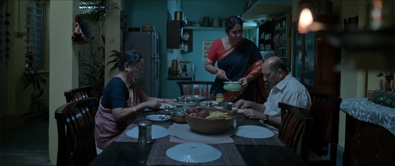 Cheekatilo (2026) movie still: medium — A domestic dining area with dark walls and a wooden table set for dinner. Three adults sit at the table…