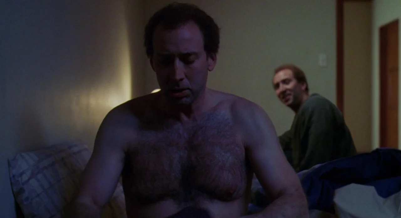Adaptation (2002) movie still: medium — A dimly lit room with blurred wall details and a bed. One man sits shirtless in the foreground while his…
