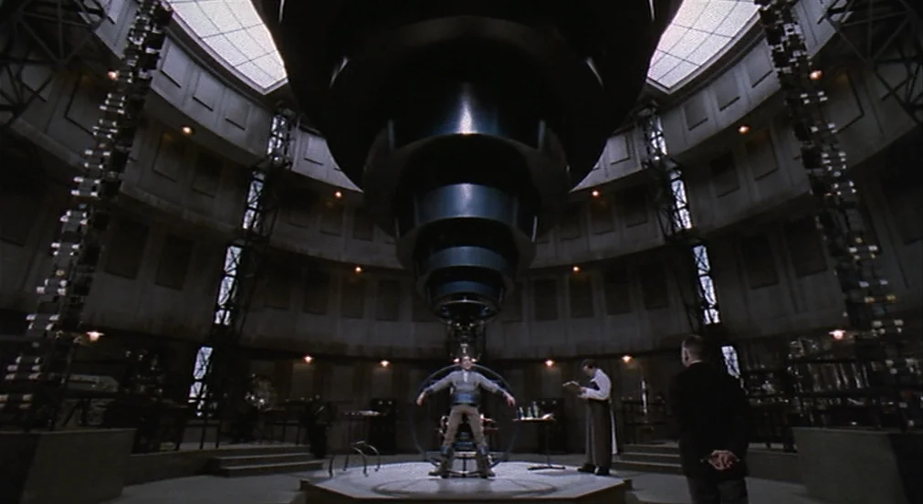 Kafka (1991) movie still: wide — A circular industrial room features a large metal structure hanging from the ceiling. A man sits strapped into a…