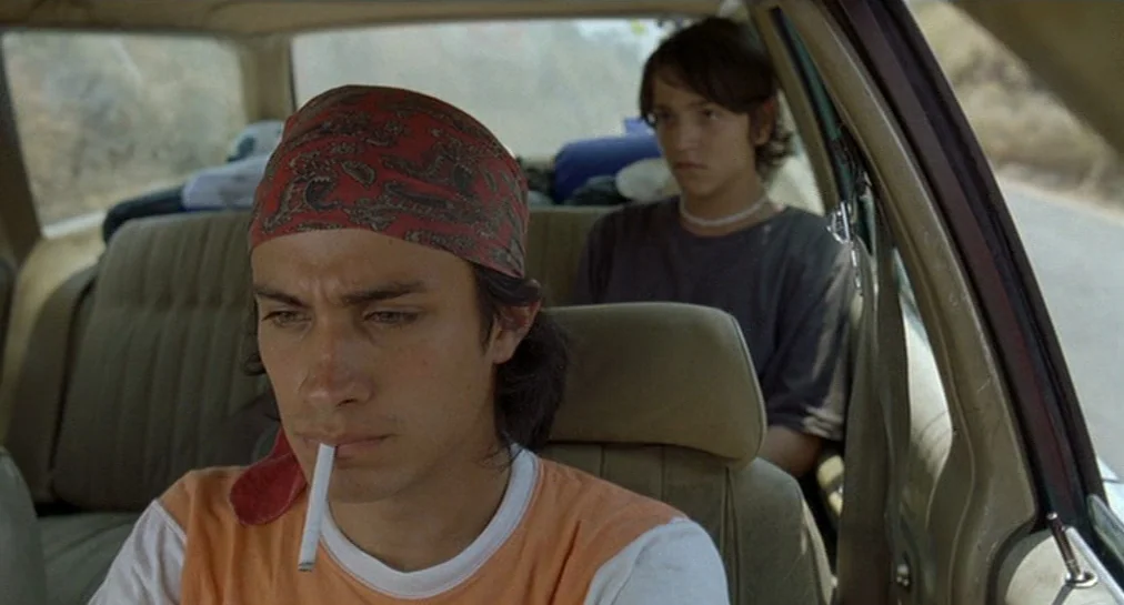 Y Tu Mamá También (2001) movie still: medium — Two men sit inside an older car with beige upholstery while traveling on road. The driver wears a…