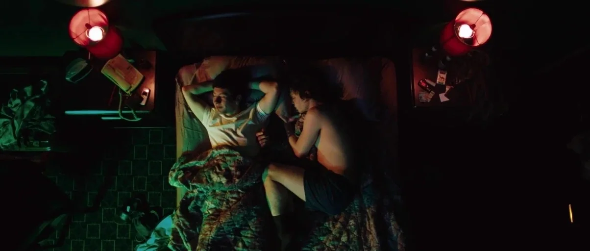 American Animals (2018) movie still: wide — A dimly lit motel room features a bed positioned directly beneath the camera. Two young men lie under a…