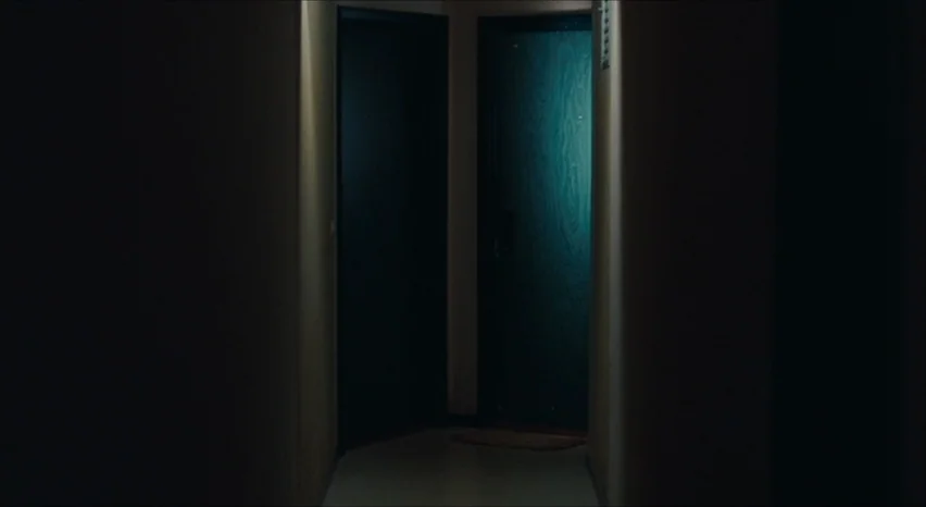 35 Shots of Rum (2008) movie still: establishing — A dimly lit narrow hallway features two closed doors set into dark walls. A teal light shines…