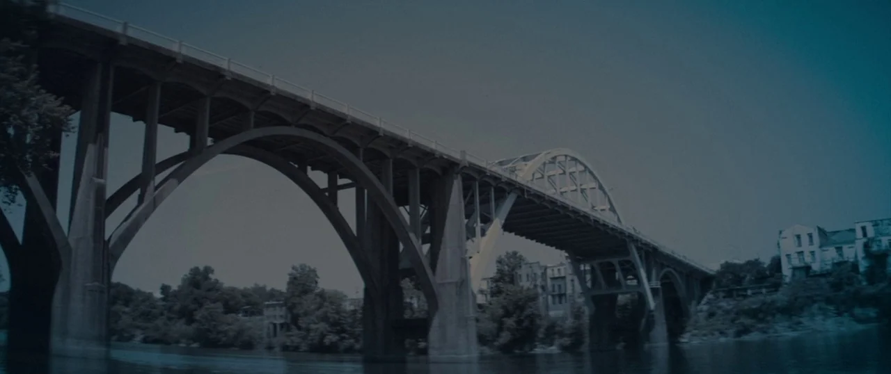 Selma (2014) movie still: establishing — A massive concrete arch bridge extends across a wide river under a cloudy sky. The structure features…