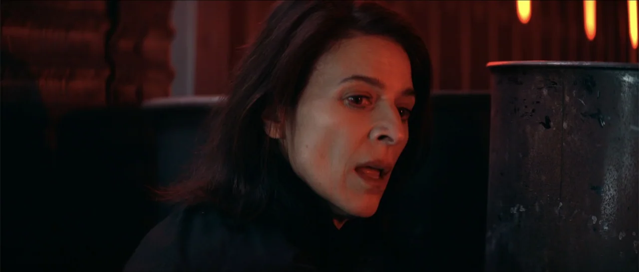 Cosmic Sin (2021) movie still: closeup — An interior industrial environment is bathed in deep shadows and harsh red illumination. A woman with dark…
