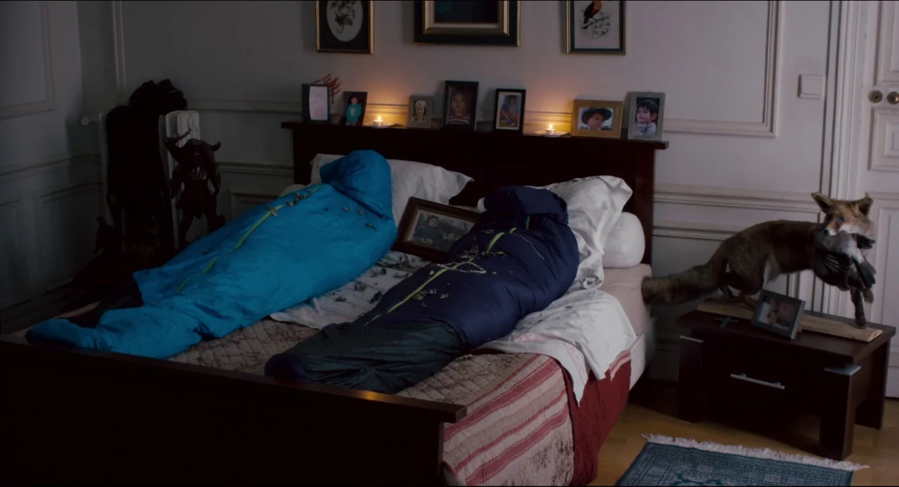 The Night Eats the World (2018) movie still: wide — A dark bedroom features a large bed occupied by two people inside sleeping bags. A taxidermy fox…