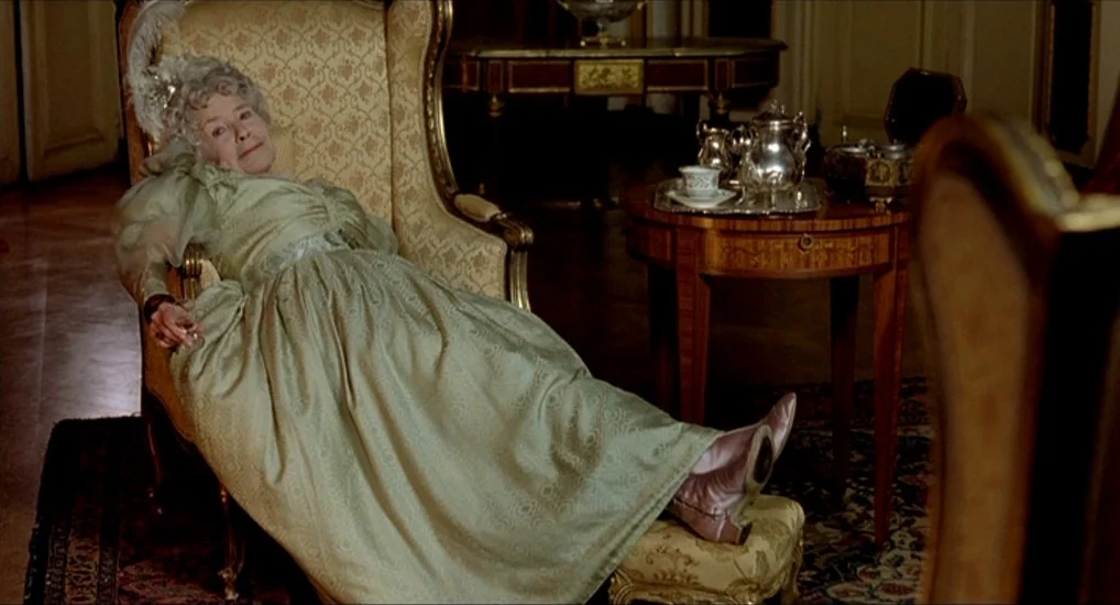 The Last Mistress (2007) movie still: medium — An opulent parlor contains an ornate wingback chair and a small mahogany tea table. An elderly woman…