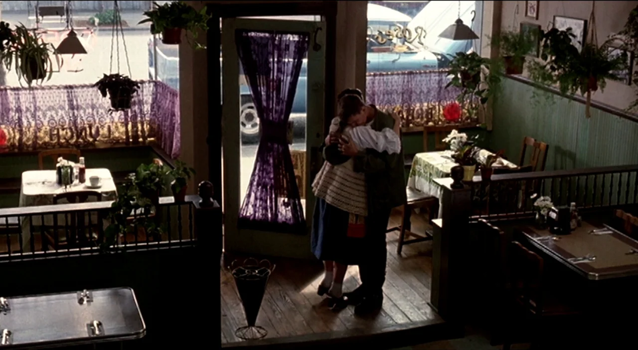 Dogfight (1991) movie still: fullbody — A diner interior features wooden flooring, small tables, and hanging plants near windows. A young man and…
