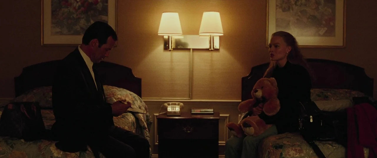 I, Tonya (2017) movie still: medium — A dated hotel room with two beds and floral bedspreads. A man sits on one bed while a woman sits on the other.