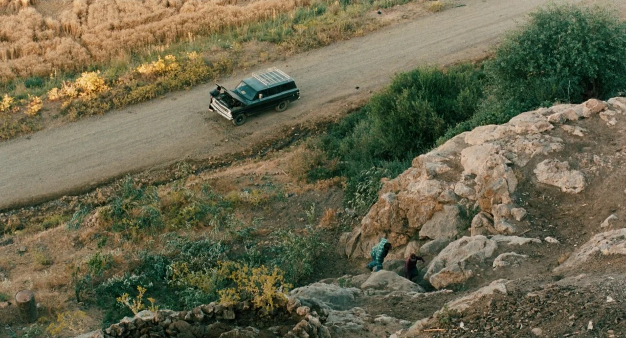 The Wind Will Carry Us (1999) movie still: wide — A rural landscape with a dirt road passing by a rocky slope. Two people walk down the hillside…