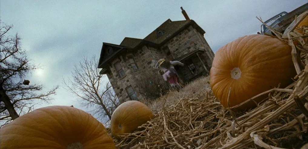 Tideland (2005) movie still: wide — A two-story stone farmhouse sits in a dry field under an overcast sky. A young child in a pink dress runs away…