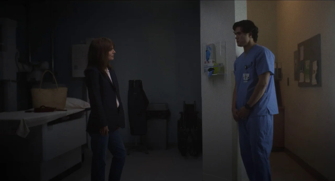 May December (2023) movie still: fullbody — A dimly lit hospital room contains a medical exam table and a wheelchair. A woman stands talking to a…