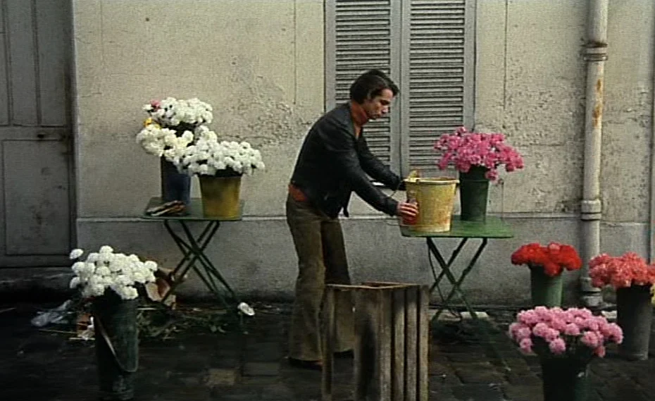 Bed & Board (1970) movie still: fullbody — A man stands outdoors near a stone building wall lined with tables of flowers. The individual reaches…